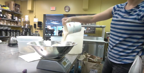 A person pours flour from a measuring cup into a mixing bowl on a kitchen scale in a bright, busy kitchen.