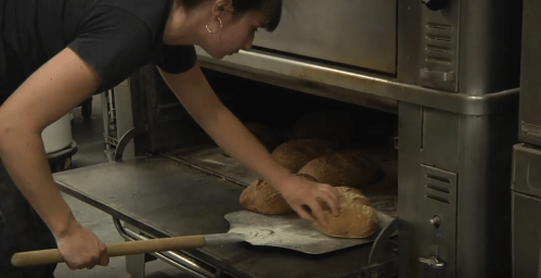 A baker removes freshly baked loaves of bread from an oven using a wooden peel.