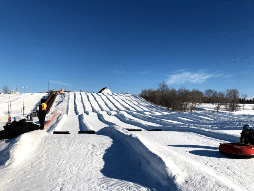 A snowy hill with multiple tubing lanes, a clear blue sky, and a few people preparing to slide down.