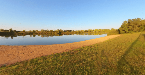 A serene lakeside view with a sandy shore, lush green grass, and clear blue skies reflecting on the calm water.