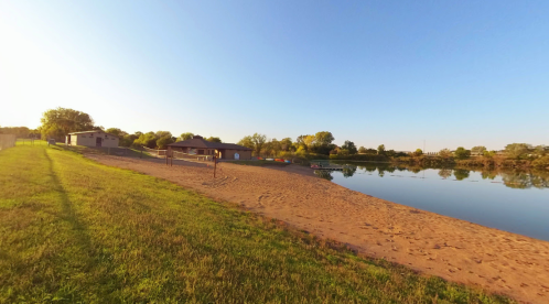 A serene lakeside scene with a sandy beach, grassy area, and a building near calm water under a clear blue sky.