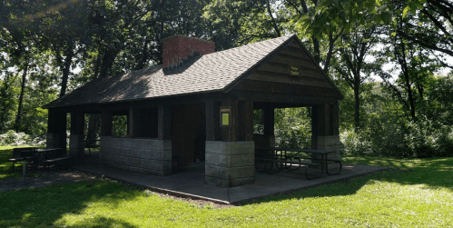 A rustic picnic shelter with a stone foundation, surrounded by trees and grass in a park setting.
