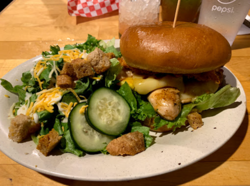 A plate with a crispy chicken sandwich, melted cheese, and a side salad with cucumbers and croutons.