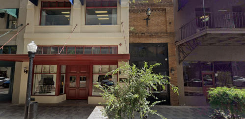 A street view of two buildings with large windows, a red awning, and greenery in the foreground.