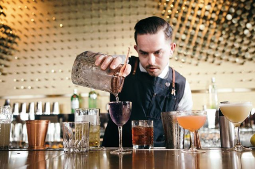 A bartender pours a colorful cocktail into a glass, surrounded by various drinks on the bar.