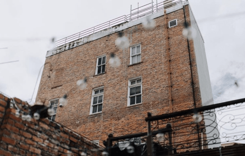 A brick building with windows, viewed from below, with string lights in the foreground and a cloudy sky above.