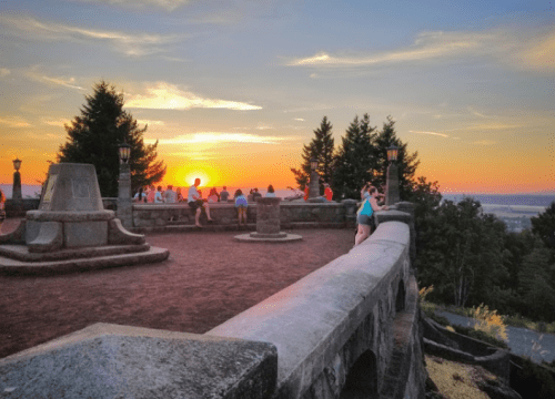 People enjoying a sunset view from a stone overlook, surrounded by trees and a scenic landscape.