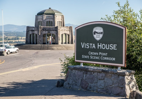 A stone building with a dome, Vista House sign in foreground, surrounded by trees and mountains in the background.