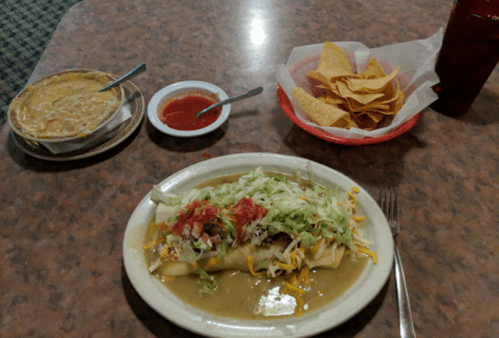 A plate with a large burrito topped with lettuce and salsa, alongside a bowl of beans, chips, and salsa on a table.