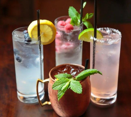 Four colorful cocktails with garnishes, including lemon, lime, and mint, served in tall glasses and a copper mug.