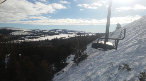 A ski lift chair hangs over a snowy landscape with mountains and trees under a bright blue sky.