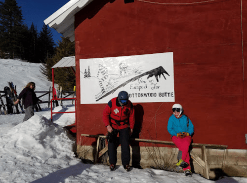 A snowy scene with a red building, a sign reading "You've Escaped to Cottonwood Butte," and two people enjoying the outdoors.