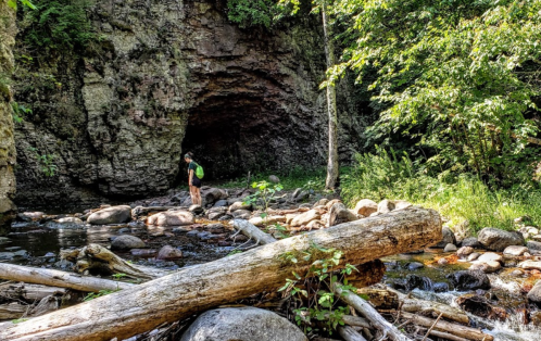 A person stands by a rocky stream near a cave entrance, surrounded by lush greenery and fallen logs.