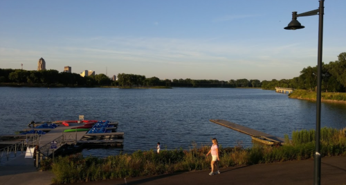 A serene river scene with a dock, colorful boats, and a person walking along the shore under a clear blue sky.