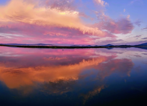 A serene landscape at sunset, featuring colorful clouds reflected in calm water with distant mountains.