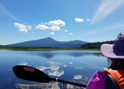 A person in a kayak views a serene lake with mountains and blue sky in the background, reflecting on the water.