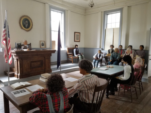 A historic classroom scene with a judge at a bench, students seated at desks, and an American flag in the background.