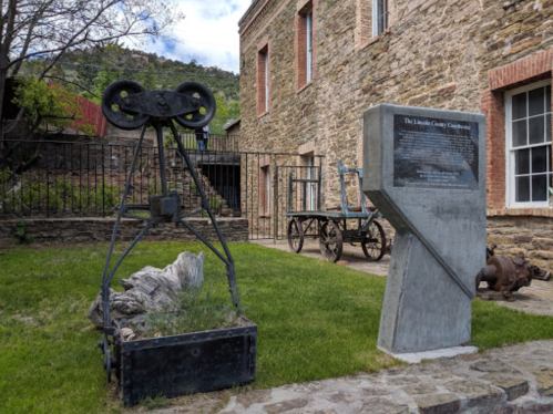 Historic site featuring a large metal pulley, stone building, and informational plaque in a grassy area.