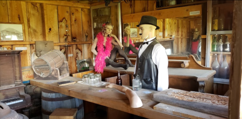 A vintage bar scene with a mannequin in a tuxedo and a woman in a pink feather boa, surrounded by wooden decor and bottles.