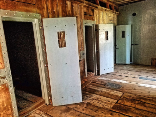Interior of a historic building featuring three open jail cells with metal doors and wooden floors.