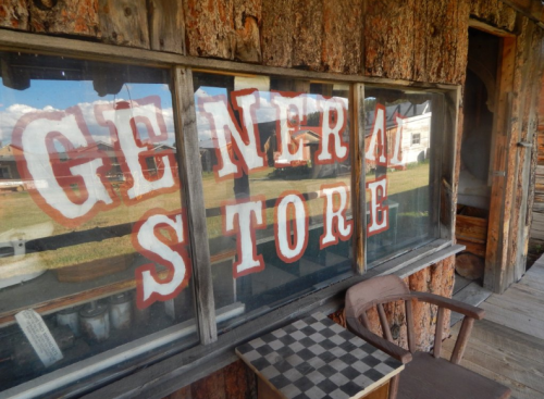 Window of a rustic general store with bold, vintage lettering and a wooden chair in front.