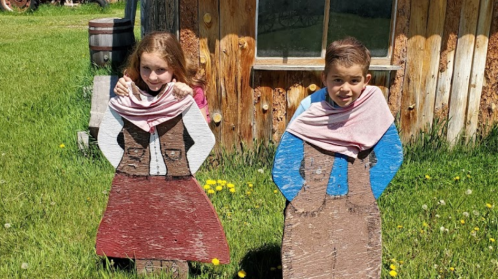 Two children pose behind wooden cutouts of people in traditional clothing, smiling in a grassy area near a rustic building.