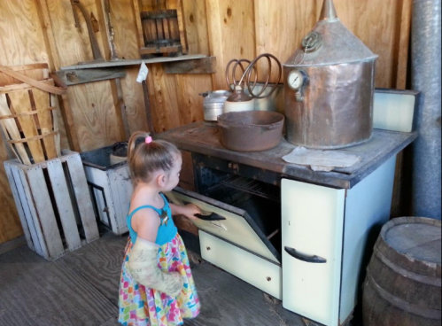 A young girl in a colorful dress explores an old-fashioned kitchen with a vintage stove and copper distilling equipment.
