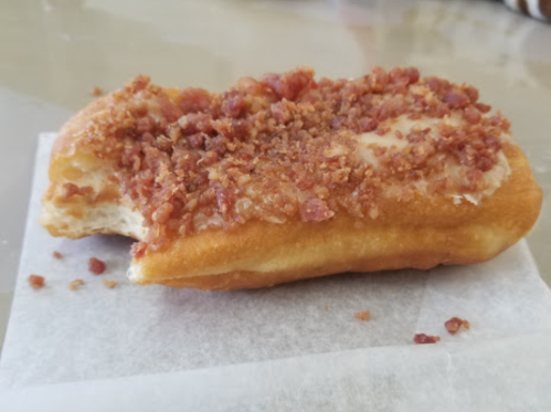 A glazed donut topped with crispy bacon bits, resting on a piece of parchment paper.