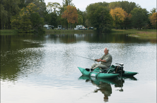 A man fishing from a green kayak on a calm lake, surrounded by trees with autumn foliage.