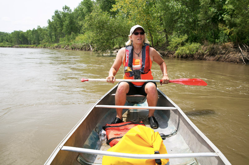 A person paddles a canoe on a river, wearing a life jacket and sunglasses, surrounded by lush greenery.
