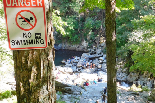 A sign warns against swimming near a rocky area with people gathered by a water pool surrounded by trees.