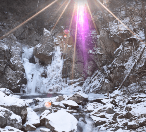 A snowy landscape featuring a waterfall, surrounded by rocky cliffs and sunlight creating a lens flare effect.