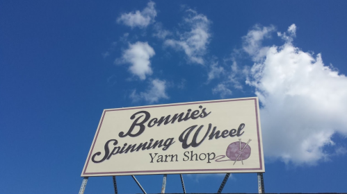 Sign for "Bonnie's Spinning Wheel Yarn Shop" against a blue sky with clouds.