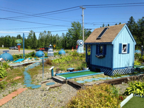 A small blue house surrounded by colorful gardens and decorative features under a clear blue sky.