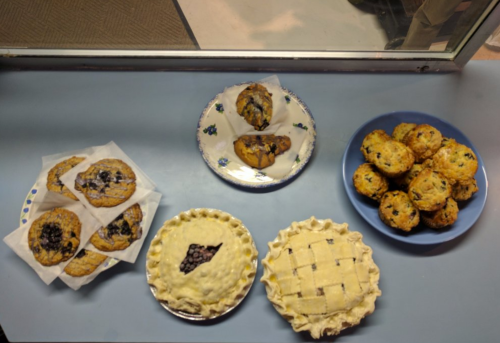 A table displaying various baked goods: cookies, muffins, and two blueberry pies.