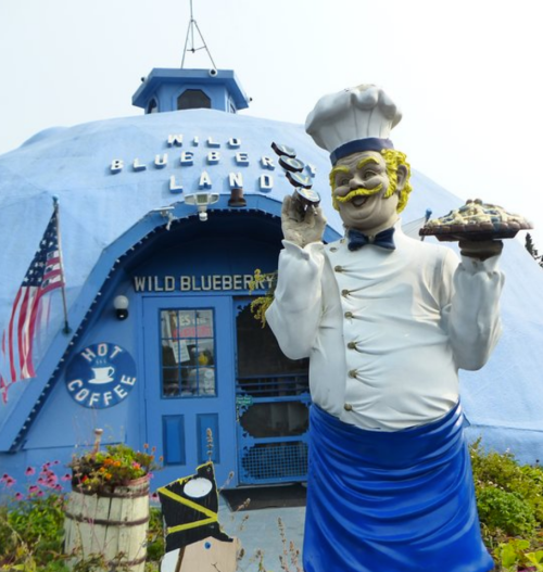 A whimsical blue building with a chef statue holding a pie, surrounded by flowers and American flags.