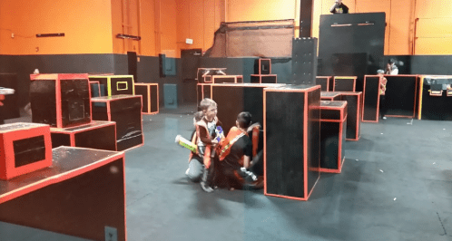 Two children in safety vests crouch behind black and orange obstacles in a laser tag arena.