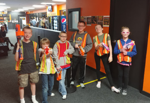 A group of six kids in safety vests holding toy blasters, smiling in a brightly lit indoor venue.