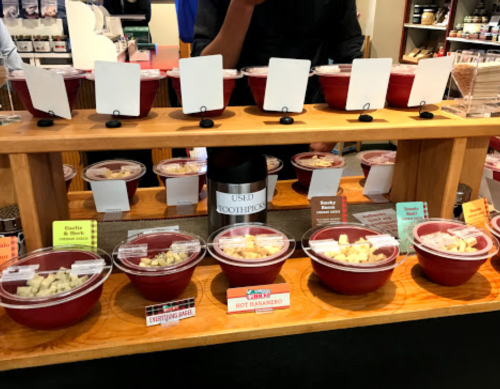 A display of various sauces in red bowls, labeled with colorful tags, on a wooden shelf with a bottle of used toothpicks.