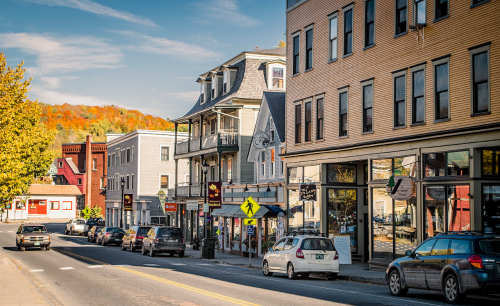 A charming street lined with shops and parked cars, framed by colorful autumn trees in the background.