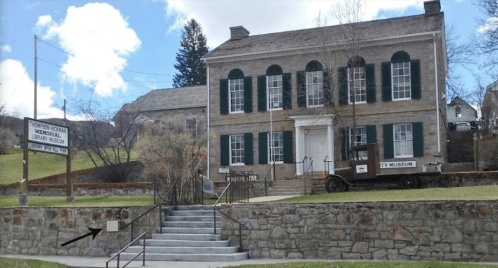 Historic stone building with green shutters, featuring a library and museum sign, surrounded by grass and trees.