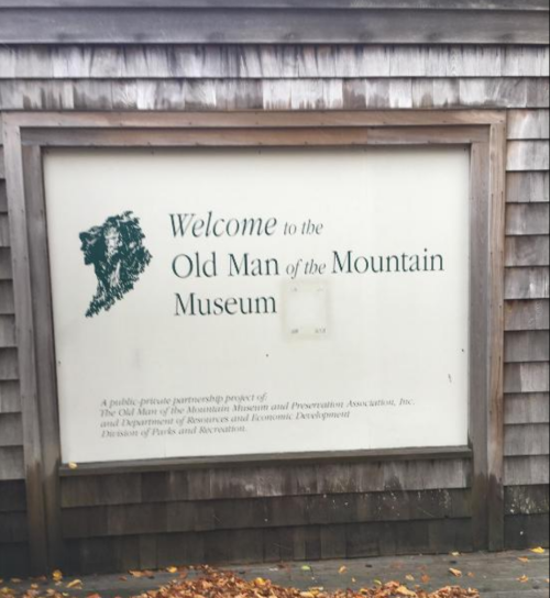 Sign for the Old Man of the Mountain Museum, featuring a green graphic and text on a wooden background.