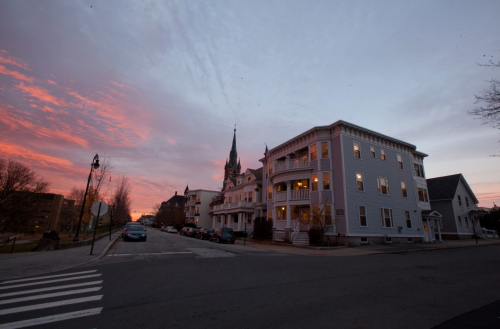 A street view at sunset featuring buildings, a church steeple, and colorful clouds in the sky.