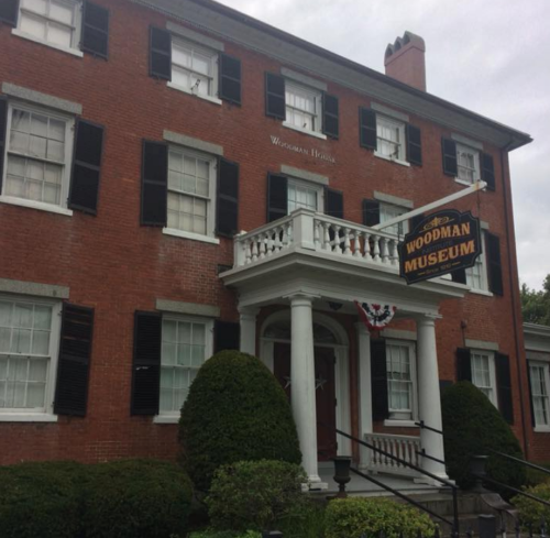 Historic red brick building with black shutters, featuring a sign for the Woodman Museum and decorative bunting.