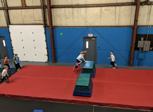 Children playing in a gym, jumping over padded obstacles on a red mat.