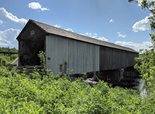 A covered bridge stands over a stream, surrounded by lush greenery and under a bright blue sky with scattered clouds.