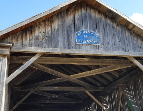 A wooden covered bridge with a sign reading "Watson Bridge 1911 Littleton ME" against a clear blue sky.