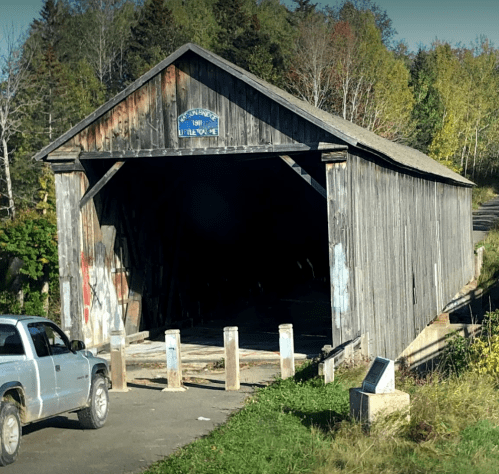 A weathered wooden covered bridge with a sign, surrounded by trees in autumn colors, and a truck parked nearby.
