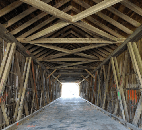 A view inside a covered bridge, showcasing wooden beams and a light source at the far end.