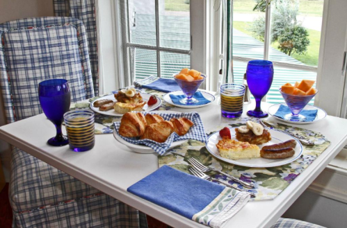 A cozy breakfast table with plates of food, blue glasses, and fruit, set by a window with a view.